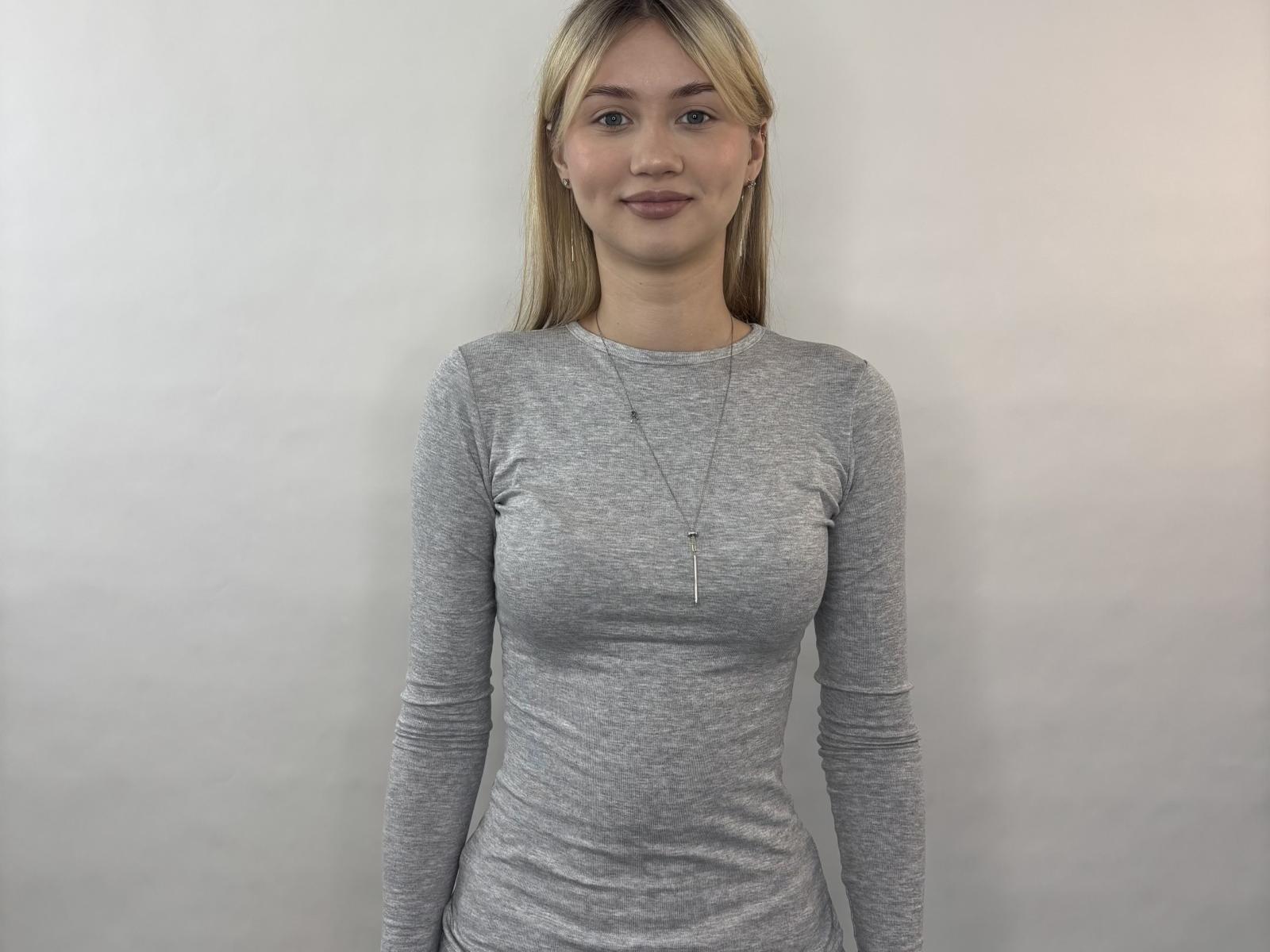 Venezia. Donna bionda, camicia a maniche lunghe grigia, seno piccolo, figura snella, in piedi, capelli lisci, sorriso discreto, collana, sfondo bianco, abbigliamento casual, pelle chiara, senza trucco..