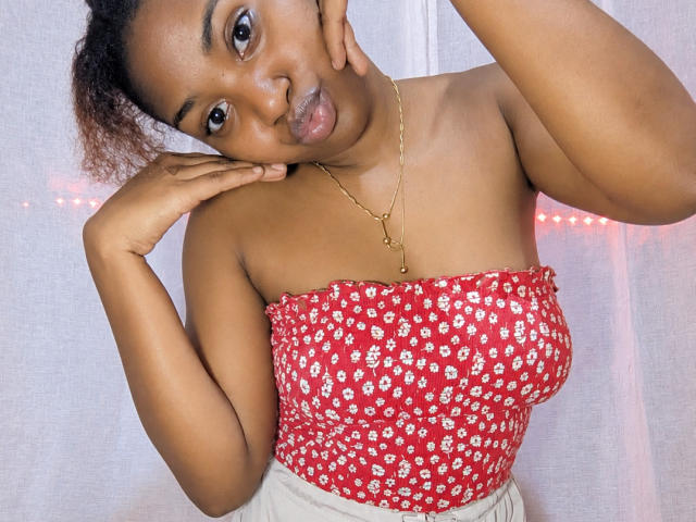 Yollande with brown skin, curly hair, wearing a red floral strapless top, gold necklace, pouty lips, hands near face, looking at camera. Her tits are small and her ass is hidden.