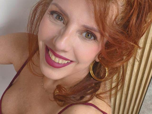 Khharha, con i capelli rossi, gli occhi verdi e il rossetto viola, sorride alla fotocamera, indossando orecchini a cerchio dorati e un reggiseno rosa, mostrando il seno..