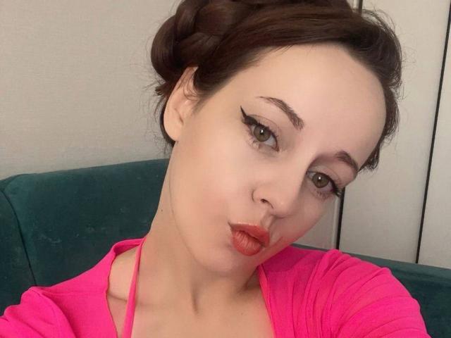 SabrinaLavv ha la pelle chiara, gli occhi verdi e il rossetto rosso, con i capelli castano scuro raccolti in una treccia, indossa una maglietta rosa e fa la smorfia con le sue labbra rosa..