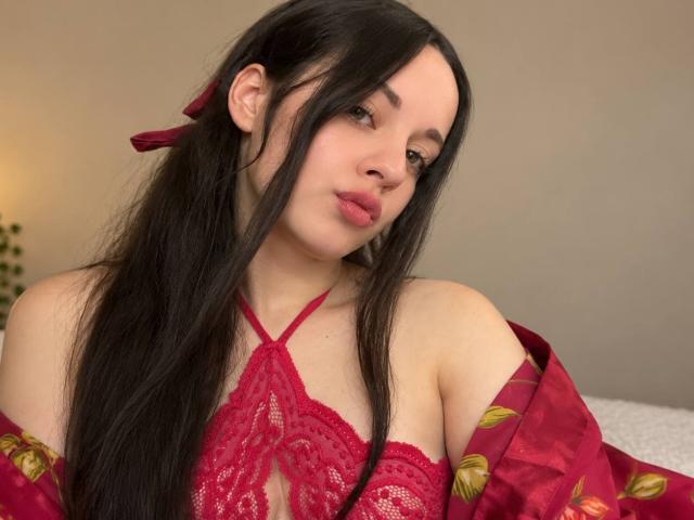 SabrinaLavv with long black hair, red lace bra, and matching robe, pouty lips, looking down, red ribbon in hair, pale skin, soft light, bedroom setting.