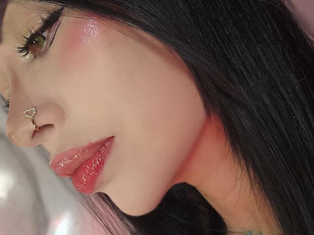 Kkole's face, showing glossy pink lips, shiny green eyes, black eyelashes, gold eyeshadow, pink blush, and a silver nose ring. Her long black hair is on the right.