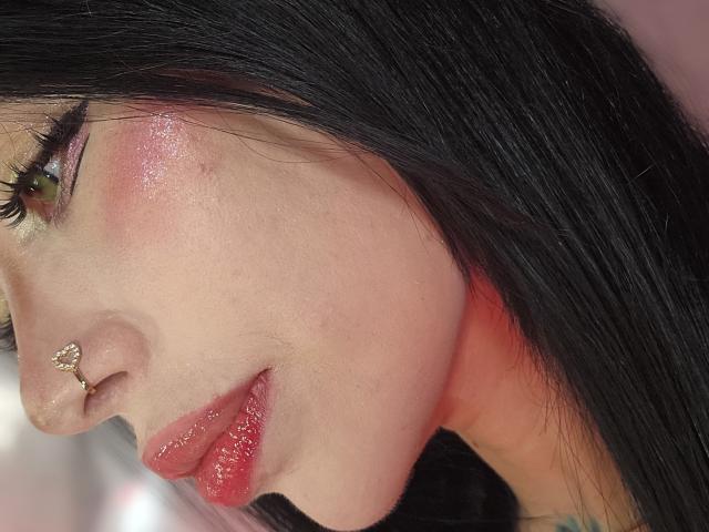 Kkole's face with shiny pink blush, green eyes, long black hair, glossy red lips, and a small nose ring. Her skin is smooth and fair.