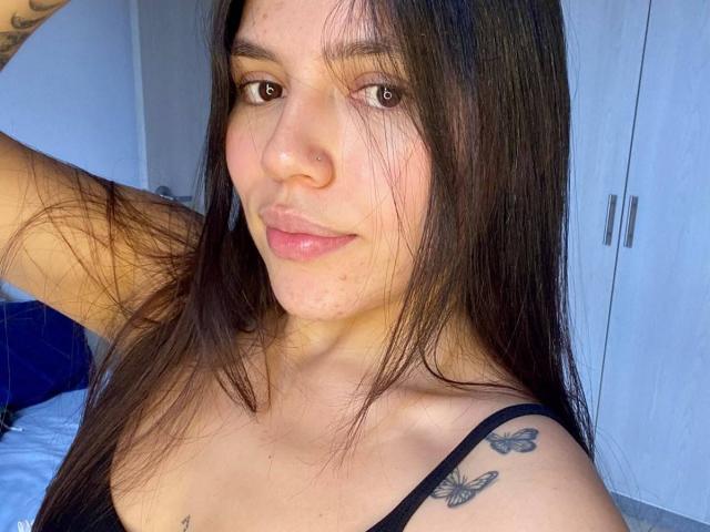 SophiaMarx with long brown hair, brown eyes, nose piercing, black strap, butterfly tattoos on shoulder, smiling, slight acne, arm raised, wearing black top, blue background.