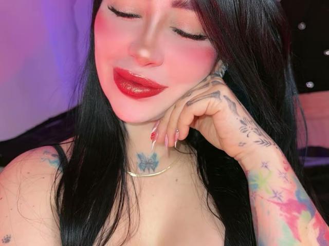 Kkole with black hair, pink blush, red lipstick, and tattoos on her neck and arm, touching her cheek, showing off her red nail polish and gold necklace.