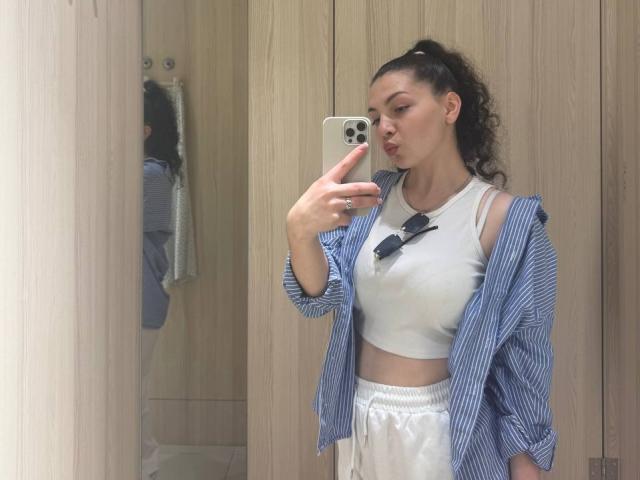 Cristal Erickson, met krullend zwart haar in een paardenstaart, draagt een blauw gestreept shirt, een witte crop top en witte broek, en maakt een selfie in de spiegel met haar iPhone..