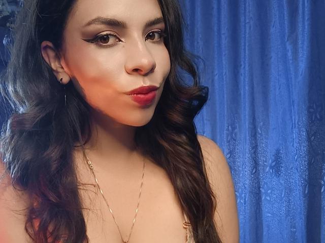 VioletaStone, avec ses longs cheveux bruns ondulés, son rouge à lèvres rouge et son eye-liner, faisant la moue avec ses lèvres rouges, portant un collier en or, devant un rideau bleu. Ses seins sont partiellement visibles..