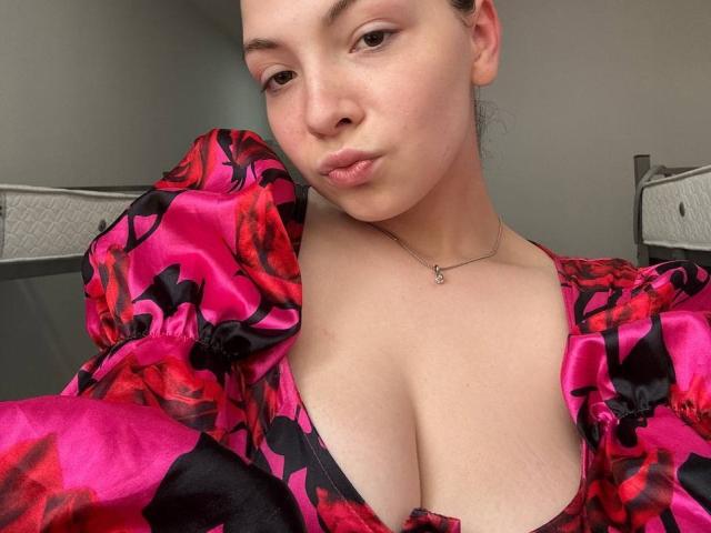 Cristal Erickson trägt ein auffälliges, pinkes und rotes Kleid mit Blumenmuster und präsentiert darin ihre Brüste, ihr schmollendes Gesicht und eine silberne Halskette auf einem Selfie. Sie hat helle Haut und braune Augen..