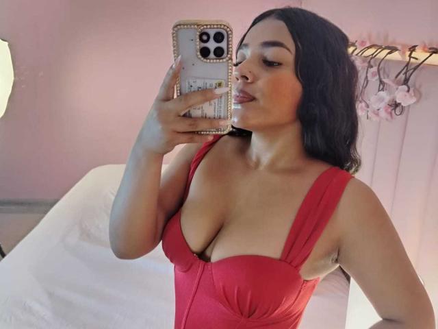 SheilaBrunette, avec de grandes seins, portant une robe rouge, se prend en photo dans un miroir, tenant un téléphone avec une coque rose, lèvres pincées, cheveux noirs ondulés, fond de mur rose..