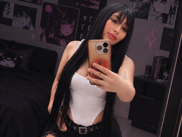 AilenRosse with long black hair, white tank top, black belt, and ripped jeans, taking a selfie in a bedroom, showing her tits, holding a phone with pink case.