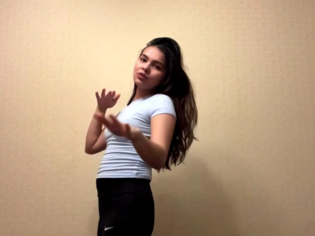 TrishMuhlbauer with long brown hair, white tight shirt, black Nike pants, standing, hands raised, slightly leaning back, looking to the side, subtle pout, soft lighting.
