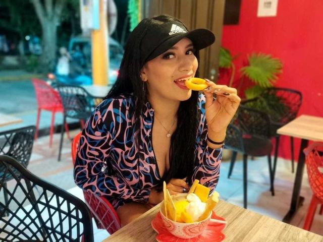 AdalinaVera, con i capelli neri, indossa un berretto nero e una camicia a righe rosa/blu e sta mangiando un frutto giallo da una ciotola di frutta su un tavolo di legno..
