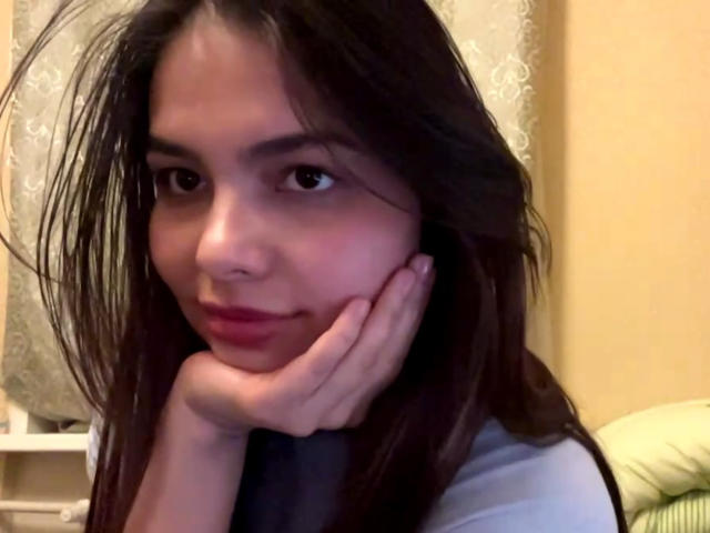 TrishMuhlbauer has long, dark brown hair, fair skin, and is resting her chin on her hand with a slight smile. Her red lips and brown eyes are visible.