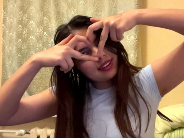 TrishMuhlbauer with long brown hair, wearing a white t-shirt, making a heart shape with her fingers in front of her face, smiling with pink lips, in a casual photo.
