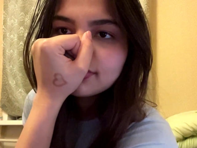 TrishMuhlbauer with long brown hair, covering her nose with her hand, has a small heart tattoo on her left index finger, wearing a light blue shirt, smiling slightly.
