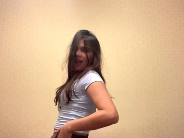 TrishMuhlbauer with long brown hair, white t-shirt, and brown pants, standing sideways, smiling with hands on her hips, exposing her ass.