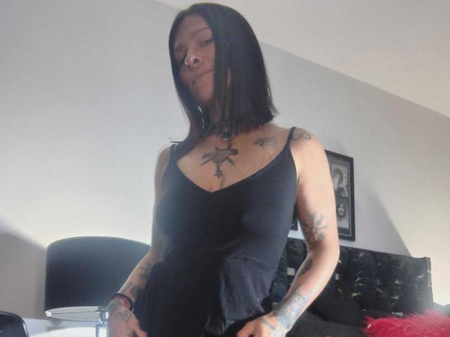 AmelieBlake with black shoulder-length hair, wearing a black tank top, showing tattoos on chest and arms, hands on hips, looking down with a slight smirk.
