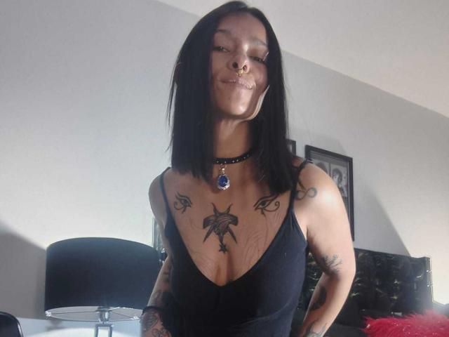 AmelieBlake with black hair, nose ring, and tattoos, wearing a low-cut black top, showing her tits, and a blue pendant, smiling slightly. She's standing indoors.