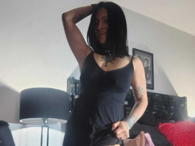 AmelieBlake with black hair, wearing a black tank top and black pants, stands with one arm raised, showing tattoos, and a hand on her waist, in a dimly lit room.