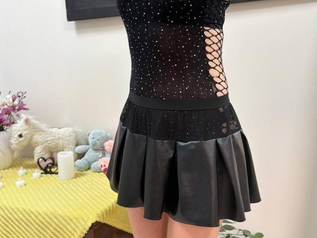 Rorria vestindo uma blusa preta transparente com brilhos e um recorte de meia-calça na lateral, combinada com uma saia de couro preta. Brinquedos de pelúcia em uma mesa amarela..