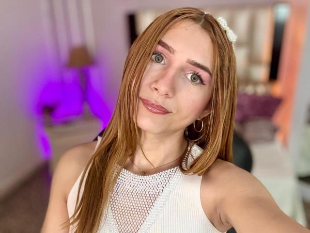 AndyDupont has long, straight, red hair, green eyes, and is wearing a white, mesh, sleeveless top, with hoop earrings, and pink lipstick, smiling softly at the camera.