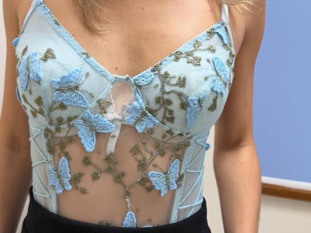 A blusa de renda azul clara e transparente de Rorria, com estampas bordadas de borboletas e detalhes em verde, revela seus pequenos seios e pele clara. Ela está vestindo calças pretas..