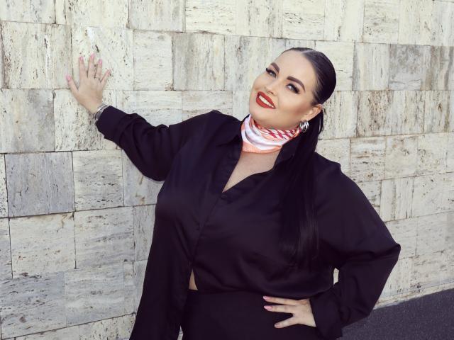 GeorgiaLerox, con i capelli neri pettinati all'indietro, rossetto rosso, camicia nera e sciarpa a righe bianche, con una mano appoggiata al muro, sorride, mostrando il seno e i glutei nella foto..