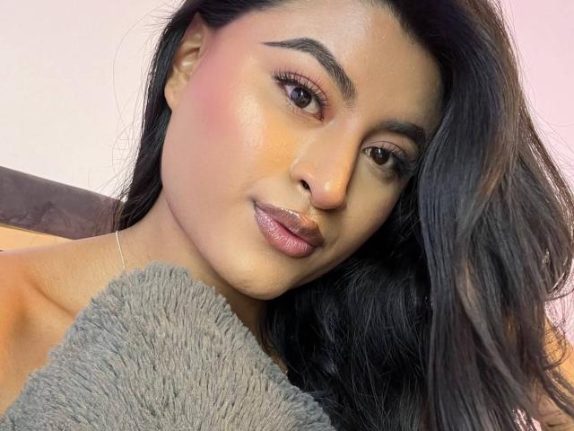 Michelle Narvaez, com cabelos escuros e ondulados, pele clara e lábios rosados brilhantes, segurando um travesseiro cinza fofo, olhando para a câmera com um leve sorriso..