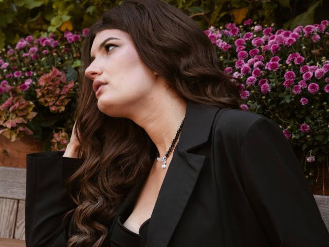 LauraLee69 with long wavy brown hair, wearing a black blazer and necklace, looking to the side with a pensive expression, surrounded by pink flowers. Her tits are partially visible.