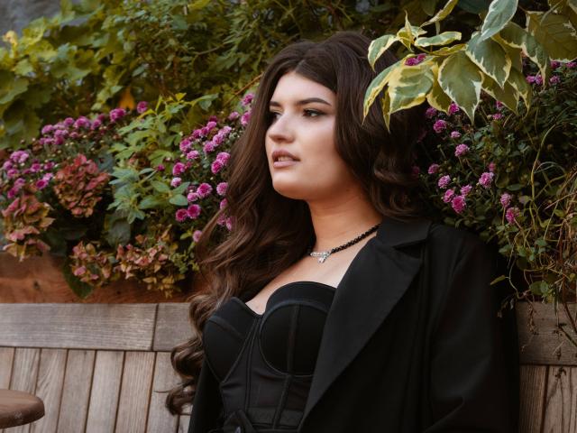 LauraLee69 has long brown hair, wearing a black dress with a strapless bust and black blazer, black necklace, looking to the side, with pink flowers and green leaves behind.