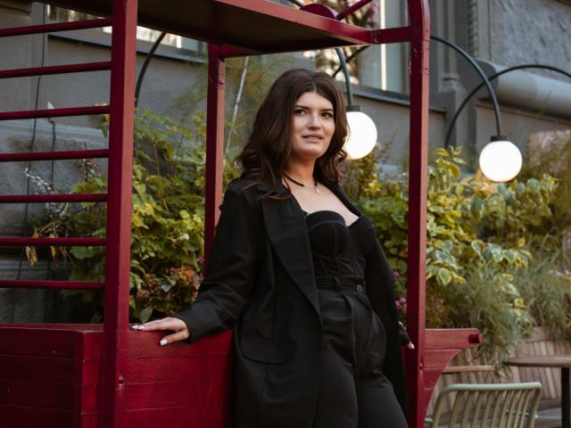 LauraLee69 with wavy brown hair, black blazer, and strapless black top, leaning on red metal structure, smiling, hands in pockets, outside at night with streetlights in background.