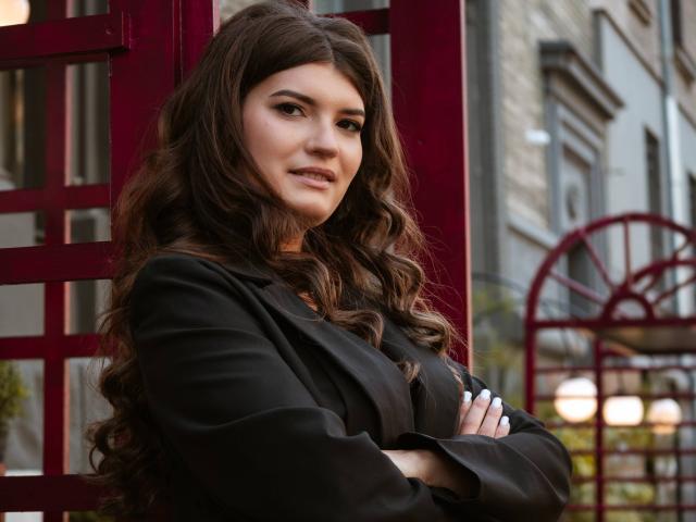 LauraLee69 with long, wavy brown hair, wearing a black blazer, arms crossed, standing in front of a red metal structure, smiling slightly, with a confident look.