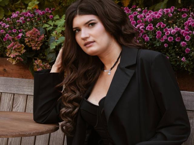 LauraLee69 with long, wavy brown hair, wearing a black blazer over a low-cut top, showing some tits, and a black necklace with a star pendant, sitting outside.