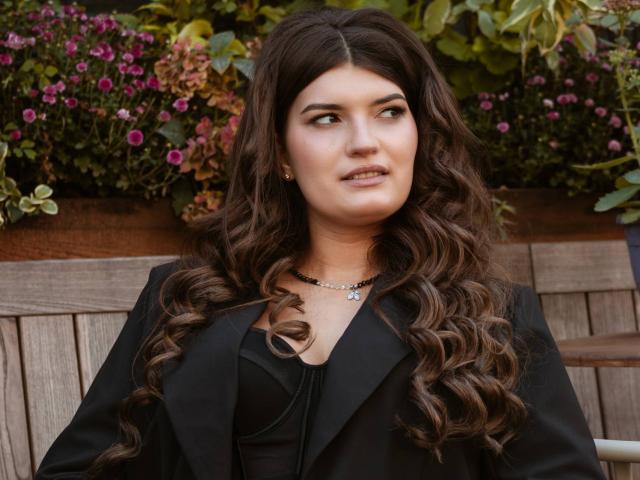 LauraLee69 with long, wavy brown hair, wearing a black blazer and necklace, looking to the side with a slight smile, showcasing her subtle makeup and small earrings.