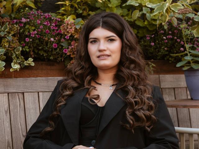 LauraLee69 with curly brown hair, black blazer, and necklace, looking at camera, wooden fence in background, small purple flowers, subtle smile, small nose ring, medium tits.