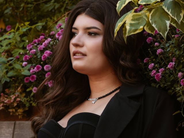 LauraLee69 has long brown hair, a black necklace, and is wearing a black dress with a strap. Her tits are visible, with a black blazer on top.