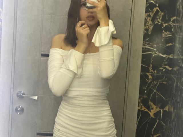 ShanekaKonig with medium tits, wearing an off-shoulder white dress, holding a phone in a mirror selfie, dark brown hair, pouty lips, in a bathroom.