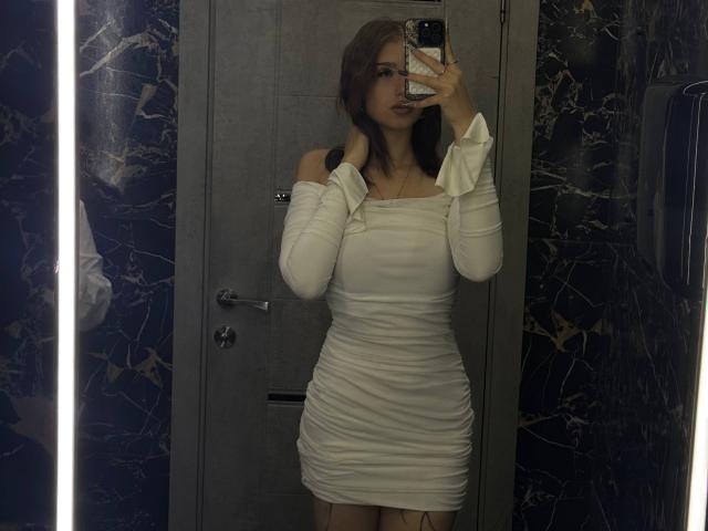ShanekaKonig trägt ein enges, weißes Kleid mit offener Schulter und Rüschen, hält ein Telefon und zeigt ihre Brüste und ihren Hintern, mit langen braunen Haaren, in einem dunklen Raum mit Marmorwänden..