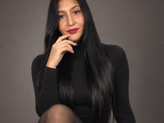 AllisonGag with long black hair, wearing a black turtleneck, has red lipstick and is touching her chin with her right hand, looking at the camera. Her pussy is not visible.