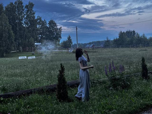 ShanekaKonig se tient dans un champ herbeux, expirant de la fumée, vêtue d'une longue robe grise, ornée de fleurs violettes, avec une bûche tombée au premier plan, sous un ciel bleu nuageux..