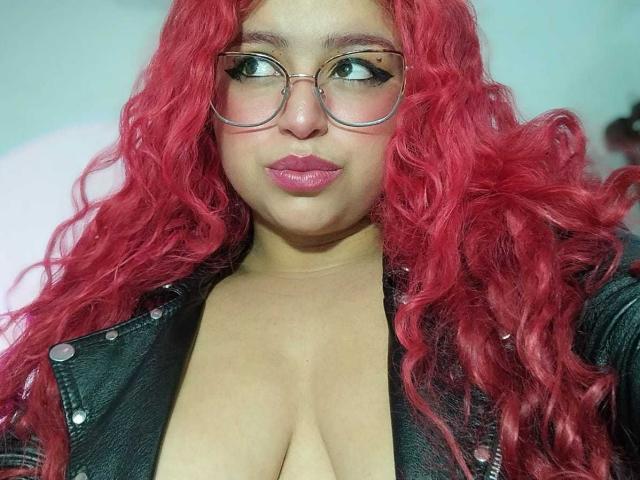 Estefy69 con cabello rizado rojo, gafas, lápiz labial rosa y una chaqueta de cuero negro que muestra sus grandes, redondos y voluptuosos senos. Está mirando hacia un lado con una ligera sonrisa..