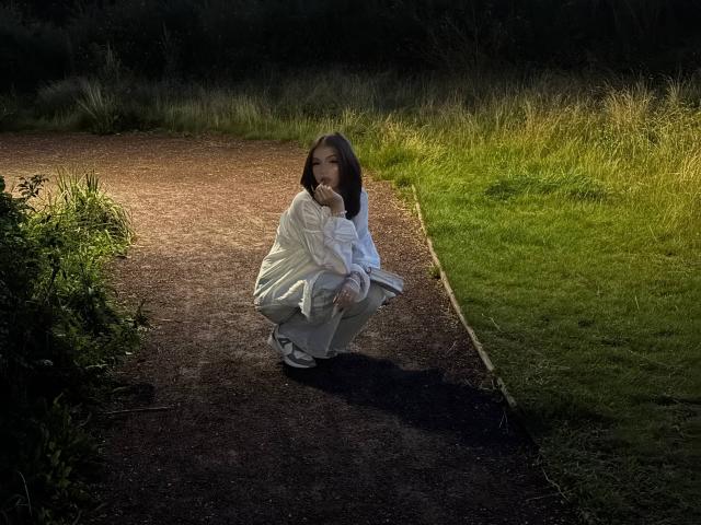 ShanekaKonig sitzt auf einem dunklen Pfad, trägt eine weiße Kapuzenjacke und graue Hosen, ihre Hand ist in der Nähe ihres Mundes, und sie befindet sich in einem grasbewachsenen, sonnendurchfluteten Bereich..
