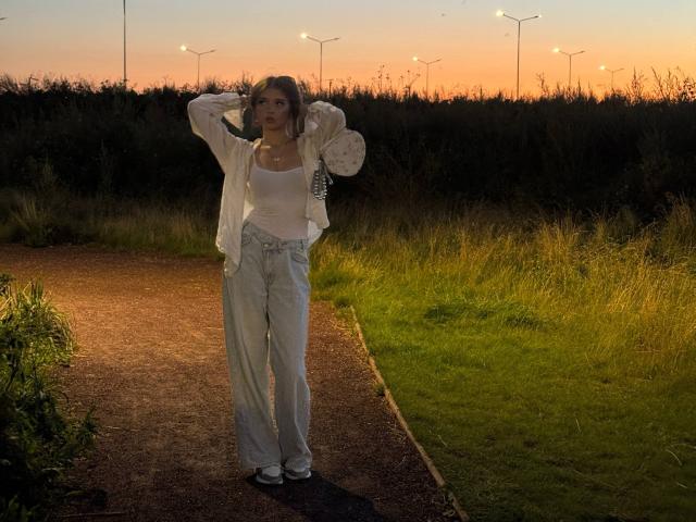 ShanekaKonig stands on a gravel path at sunset, wearing a white tank top, light jeans, and a white jacket, with hands behind her head, looking to the side.