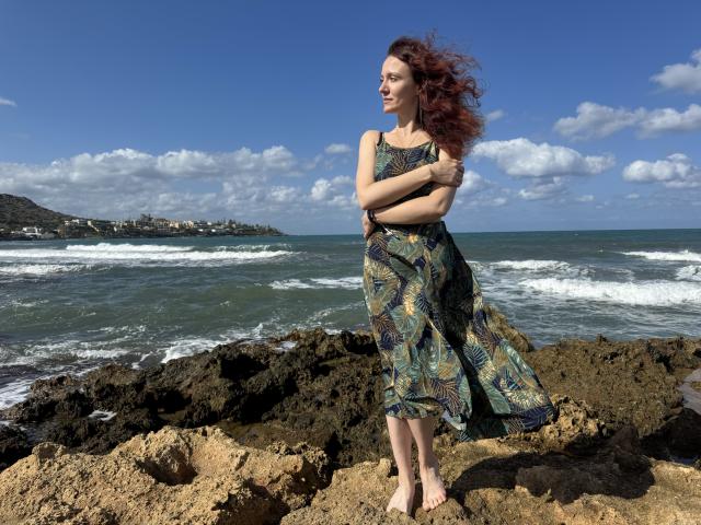 BarbaraStily debout sur une plage rocheuse, portant une robe fleurie, les bras croisés, les cheveux roux flottant au vent, pieds nus, avec des vagues de l'océan en arrière-plan, un ciel bleu clair avec des nuages ​​duveteux..