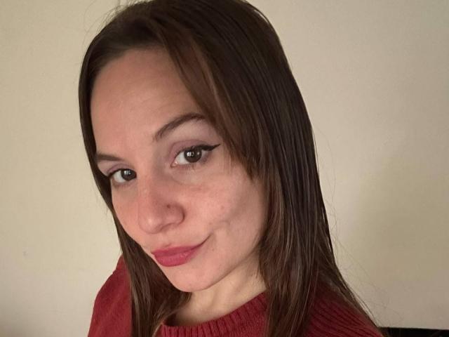RomiSweety con capelli castani, pelle chiara, rossetto rosa, un leggero trucco per gli occhi e un maglione rosso, sorride leggermente, mostrando un viso carino e un nasino piccolo..