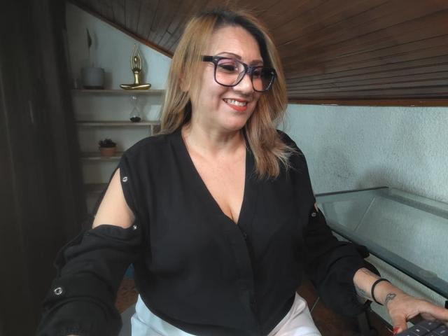 MatureYayita with glasses, black V-neck shirt, showing some cleavage, and white pants, smiling, blonde hair, wooden ceiling, shelf with golden award, and small plant in background.