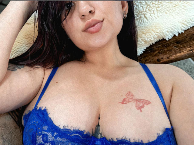 Mariana Rusell mit dunkelroten Haaren, einem blauen Spitzen-BH und einem Schmetterlingstattoo auf der Brust, die ihre Brüste zeigt, mit einem leichten Lächeln und der Hand im Haar..