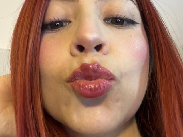 Het gezicht van MiaaRouge, met volrode, licht glanzende lippen, doet een kusgezicht, rood haar, lichte huid, donkere ogen met lange wimpers, lichte blos op de wangen, close-up selfie..