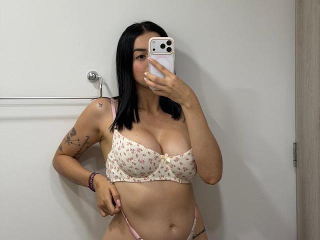IsabellaJames with black hair, wearing a floral bra and pink thong, showing her big tits and pulling down her thong, holding a phone in a bathroom mirror selfie.
