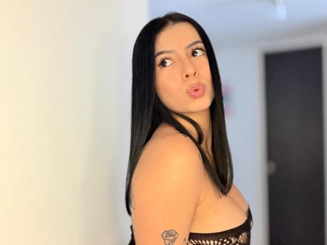 IsabellaJames with black hair, pouty lips, and a tattoo on her upper arm, wearing a strapless black lace top, showing her tits. She's looking over her shoulder.